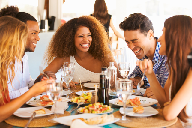 Mehrere Personen unterschiedlicher Herkunft sitzen lachend im Restaurant beim Essen und unterhalten sich.